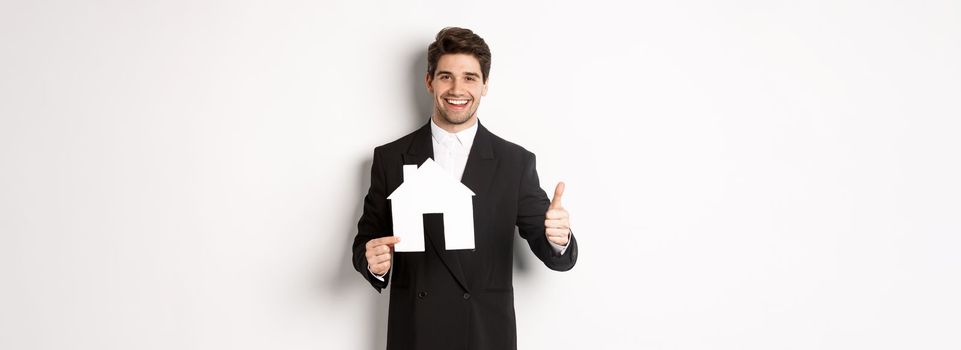 Portrait of confident real estate agent showing house maket and thumb-up, standing against white background.