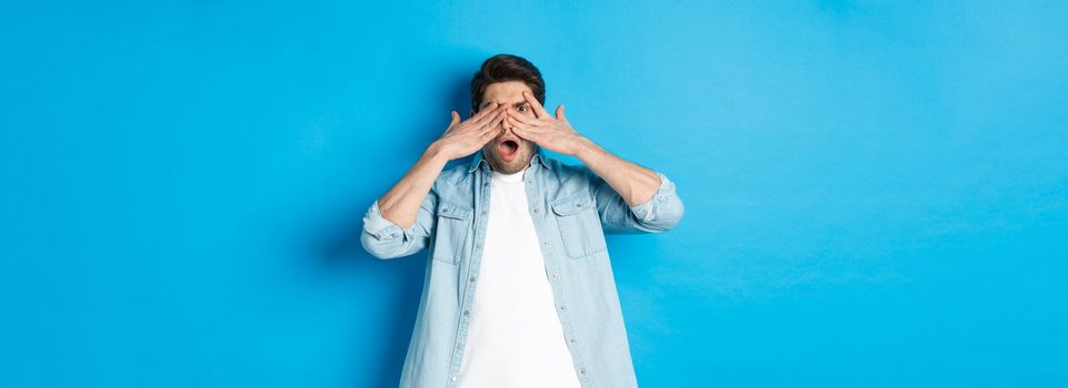 Shocked man covering eyes and peeking through fingers, stare at something embarrassing, standing against blue background.