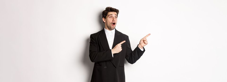 Impressed handsome man in party suit, looking at new year promo offer and pointing fingers left at banner, standing over white background.