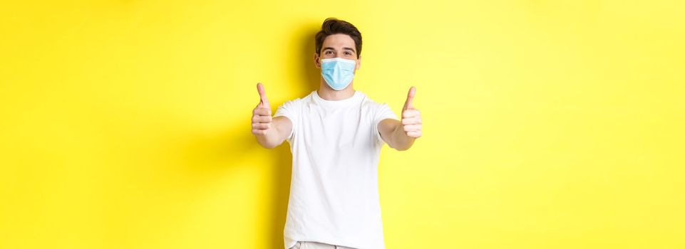 Concept of coronavirus, pandemic and social distancing. Confident man protecting himself from covid-19 with medical mask, showing thumbs up in approval, yellow background.