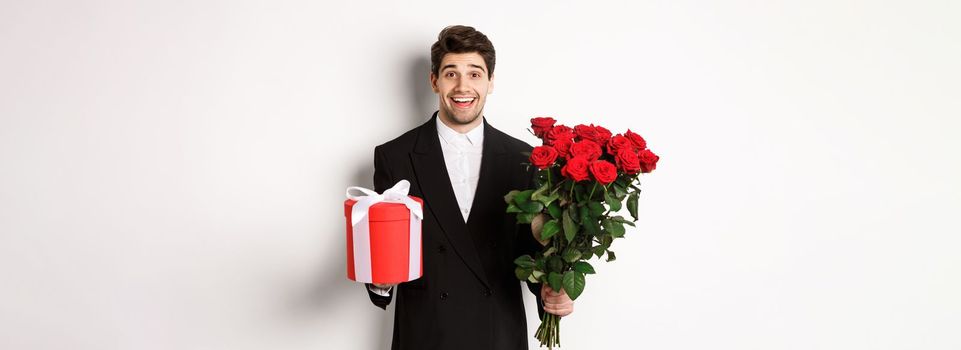 Concept of holidays, relationship and celebration. Image of handsome smiling guy in black suit, holding bouquet of red roses and giving present on new year, white background.