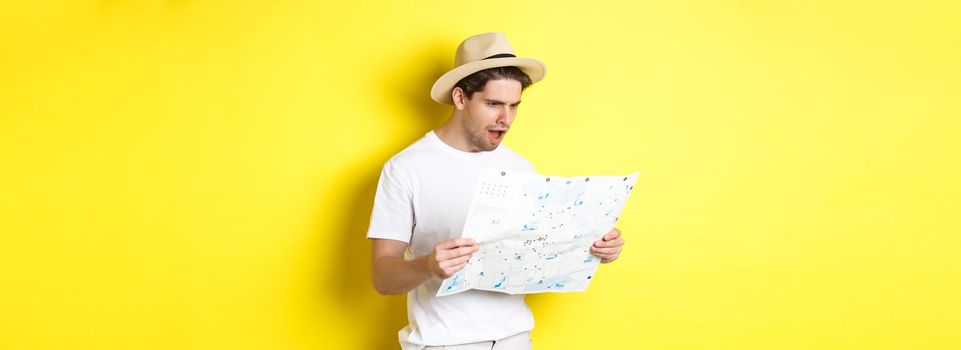 Travelling, vacation and tourism concept. Tourist looking displeased and shocked at road map, standing against yellow background.