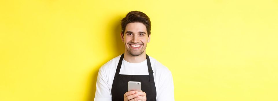 Close-up of handsome barista sending message on mobile phone, smiling happy, standing over yellow background.