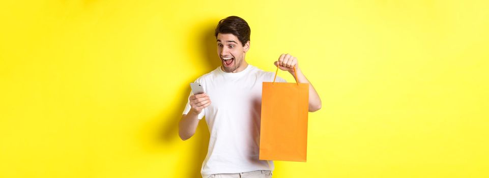 Concept of discounts, online banking and cashback. Surprised man showing shopping bag and looking happy at mobile screen, standing against yellow background.