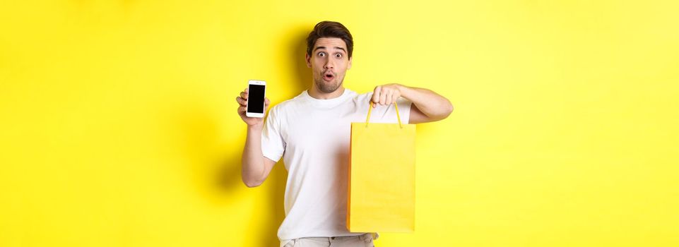 Surprised man holding shopping bag and showing smartphone screen, concept of mobile banking and app achievements, yellow background.