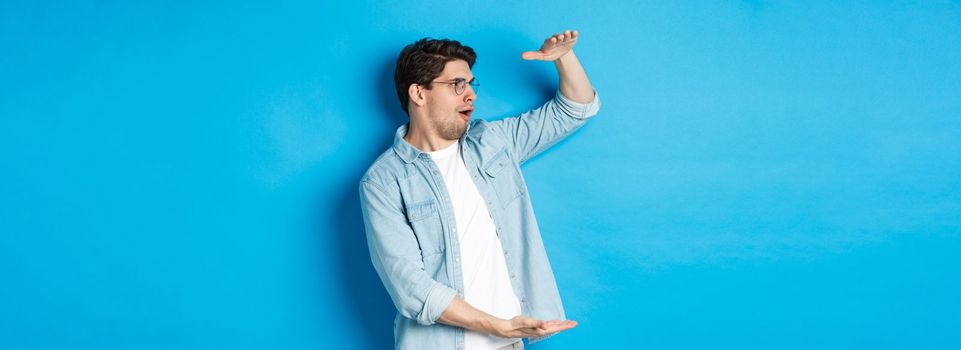 Excited handsome man showing big size object and looking amazed, standing over blue background.