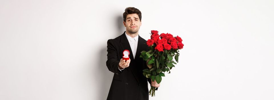 Handsome smiling man in black suit, holding roses and engagement ring, making a proposal to marry him, standing against white background.