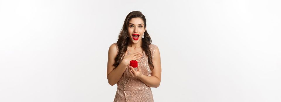 Image of amazed girlfriend holding engagement ring box, looking surprised, receive marriage proposal, wearing elegant dress, white background.