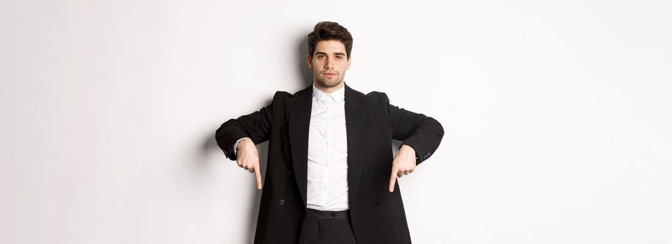 Image of handsome man dressed for formal party, wearing suit and pointing fingers down, showing advertisement or making announcement, standing over white background.