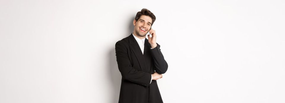 Image of handsome, successful businessman talking on phone, smiling pleased, standing in suit against white background.