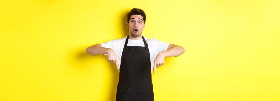 Surprised seller in black apron pointing fingers down, looking amazed at camera, standing over yellow background.