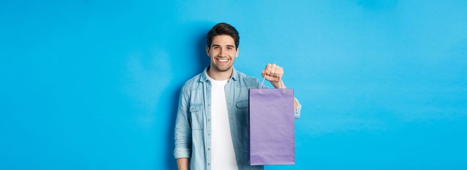 Concept of shopping, holidays and lifestyle. Young handsome man holding paper bag with present and smiling, standing over blue background.