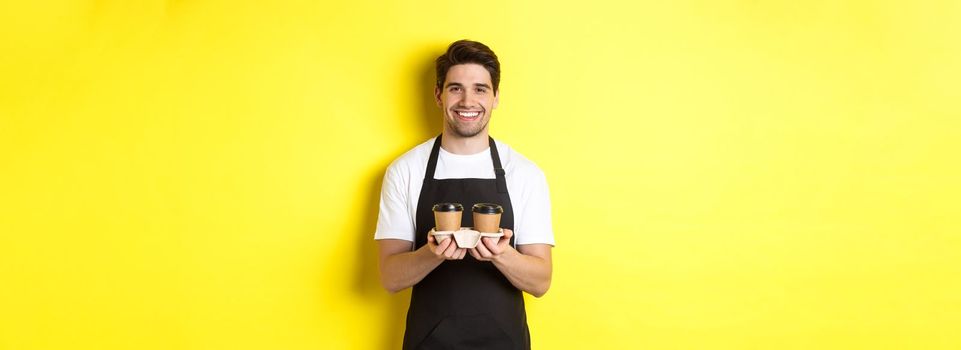 Handsome male barista serving takeaway coffee and smiling, bringing order, standing in black apron against yellow background.