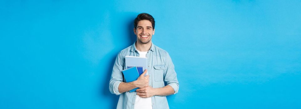 Young man holding notebooks and study material, smiling happy, standing over blue background.