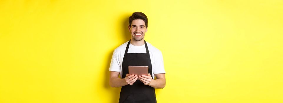 Waiter in black apron taking orders, holding digital tablet and smiling friendly, standing over yellow background.