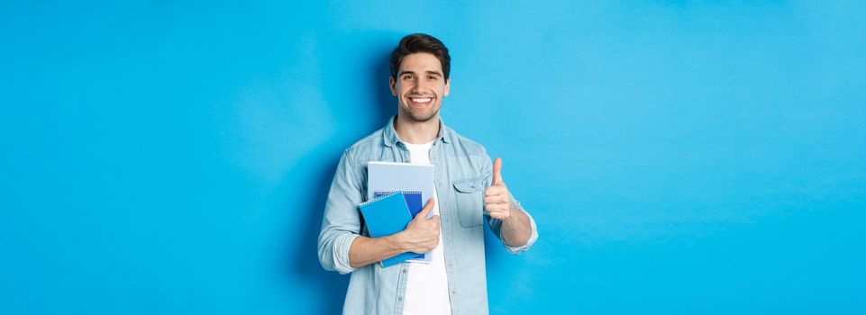 Young man student with notebooks, showing thumb up in approval, smiling satisfied, blue studio background.