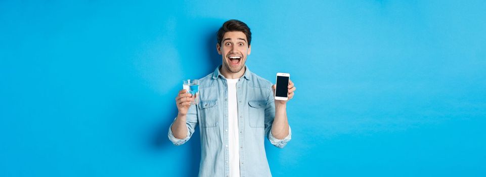 Happy man showing glass of water and mobile screen, recommending smartphone health app, standing over blue background.