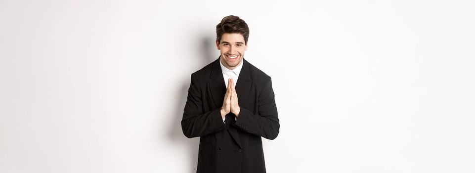 Portrait of handsome man in black suit, being grateful, saying thank you and bowing politely, smiling as holding hands together, expressing gratitude, standing over white background.