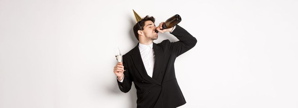 Image of handsome guy in trendy suit, drinking champagne from bottle and celebrating holiday, having a birthday party, getting drunk while standing over white background.