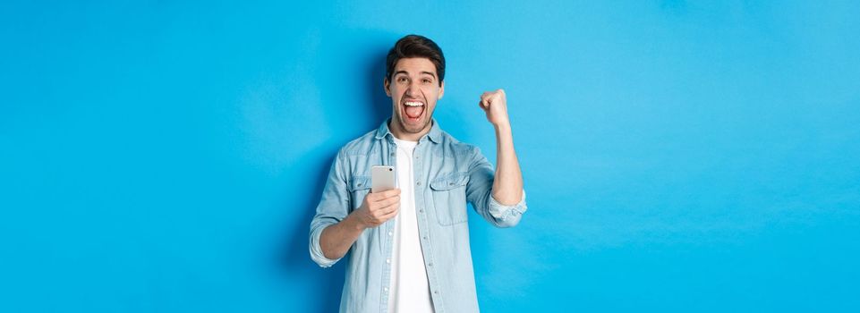 Concept of online shopping, applications and technology. Excited man shouting yes and making fist pump gesture after winning on smartphone, standing over blue background.