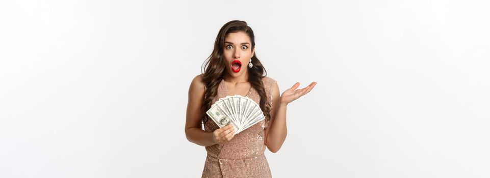 Shopping concept. Image of beautiful woman in glamour dress looking shocked, holding dollars and staring at camera, standing over white background.