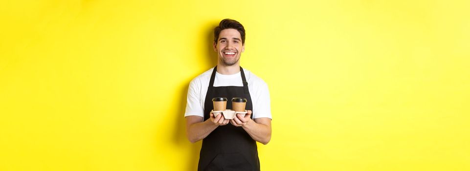 Friendly barista in black apron giving takeaway order, holding two cups of coffee and smiling, standing over yellow background.