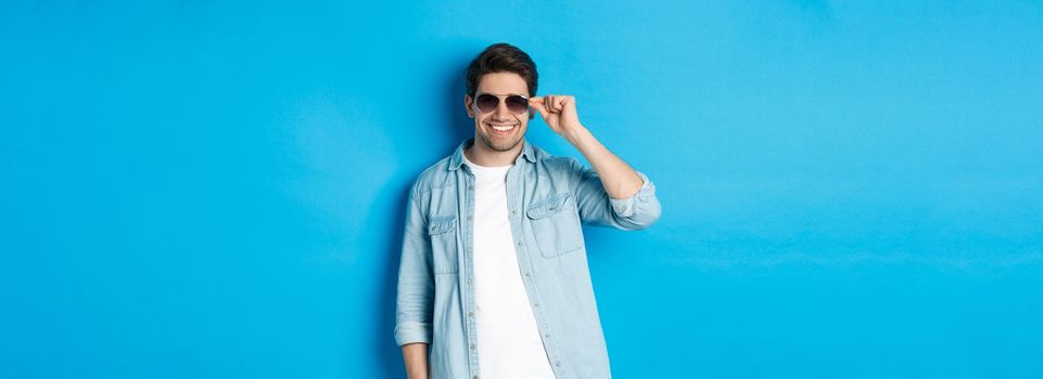 Confident macho man put on sunglasses, looking cool and sassy, standing over blue background.