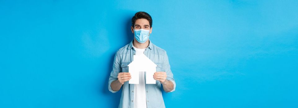 Concept of coronavirus, quarantine and social distancing. Young man searching apartment, showing house paper model, wearing medical mask, renting or buying propery, blue background.