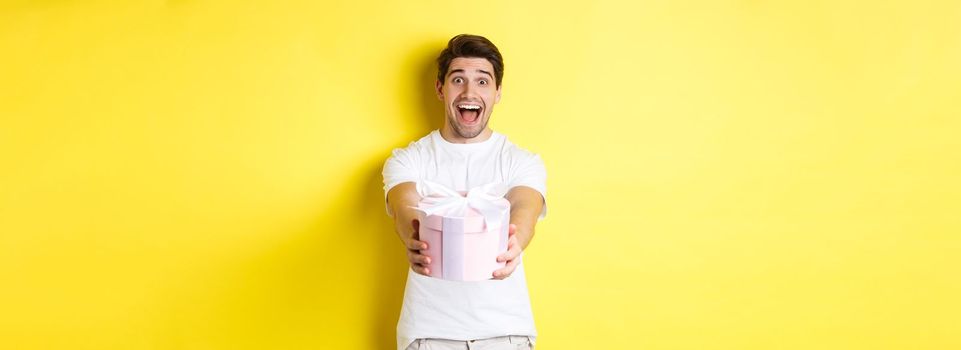 Concept of holidays and celebration. Happy man wishing happy new year, giving you gift, standing over yellow background.