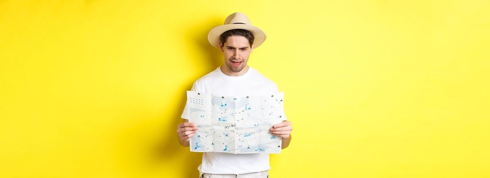 Travelling, vacation and tourism concept. Man looking confused at map during trip, cant understand, standing over yellow background.