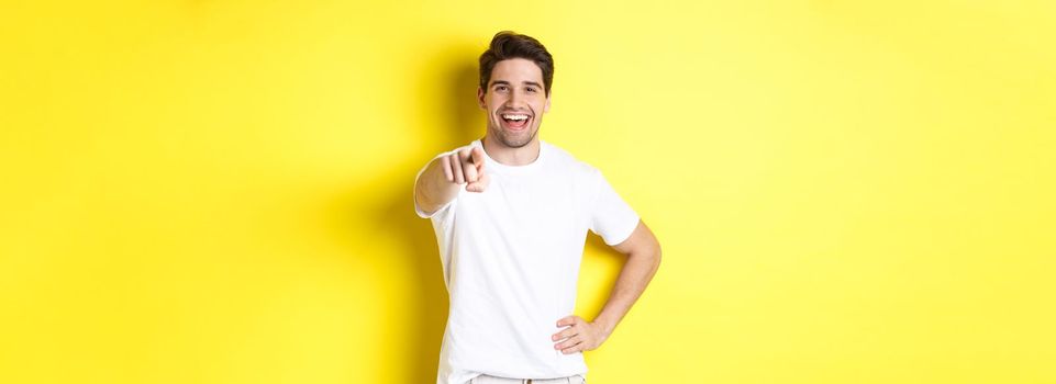 Confident smiling man pointing at you camera, standing in white clothes against yellow background.