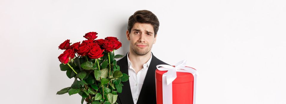 Close-up of sad man in suit, holding bouquet of red roses and a gift, standing upset against white background.