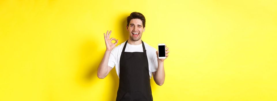 Handsome coffee shop worker showing ok sign and smartphone screen, recommending application, standing over yellow background.