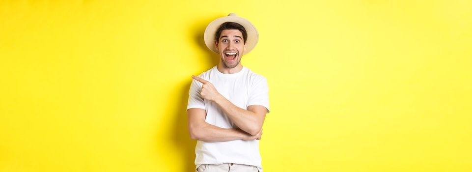 Happy male tourist in straw hat pointing finger right, showing promo offer on copy space, yellow background. Copy space