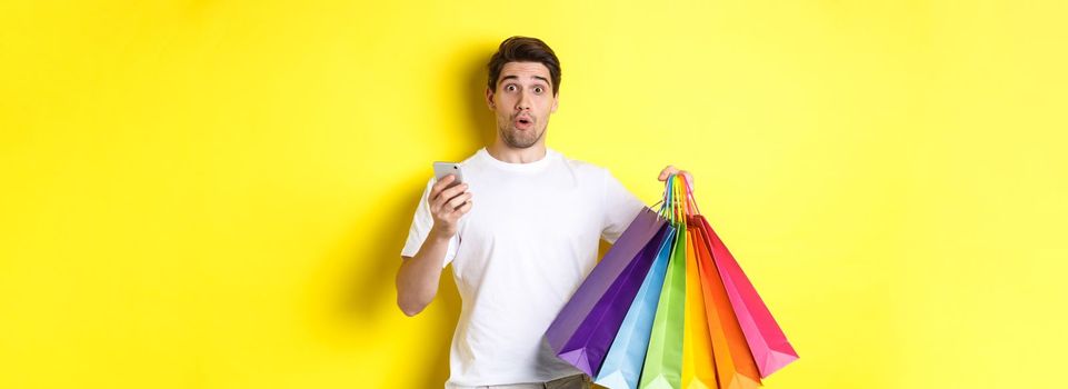 Concept of mobile banking and cashback. Surprised man holding shopping bags and smartphone, standing over yellow background.