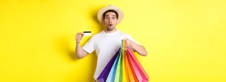Impressed man showing shopping bags with products and credit card, standing over yellow background.