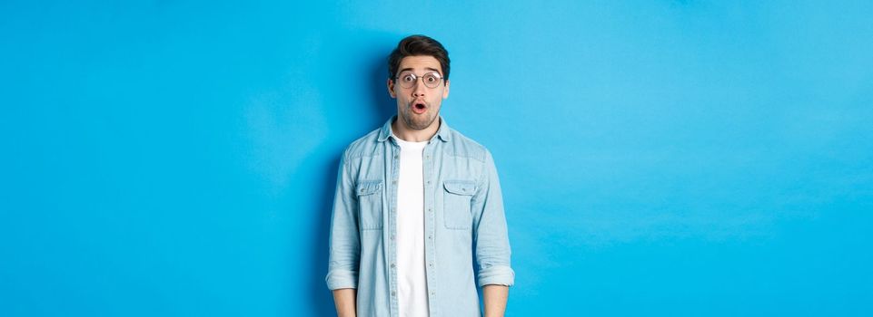 Surprised man in glasses looking at promo offer, saying wow amazed, standing over blue background.