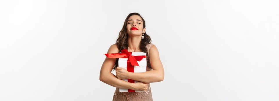 Holidays, celebration concept. Happy and thankful woman hugging Christmas gift, wearing luxury dress, receiving New Year present, standing over white background.