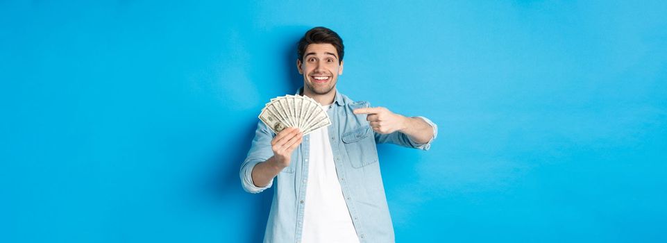Surprised smiling man in casual clothes, pointing fingers at money, standing over blue background.