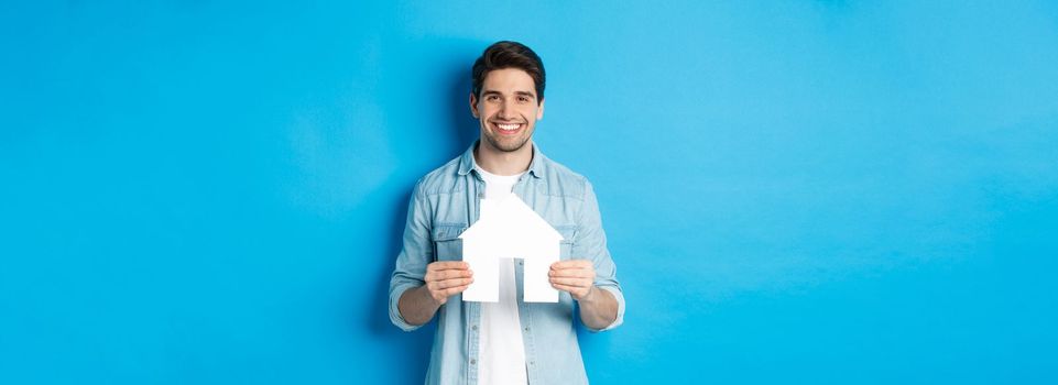 Insurance, mortgage and real estate concept. Smiling young man holding house model, searching apartment for rent, standing against blue background.