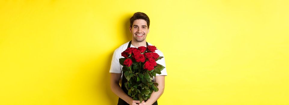 Smiling florist in black apron holding flowers, selling bouquet of roses, standing over yellow background.