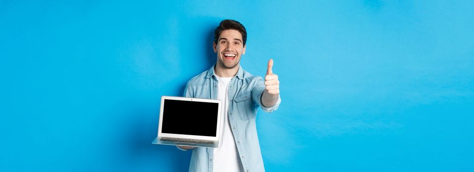 Image of satisfied handsome man showing laptop screen, thumbs-up in approval, like website or internet, standing over blue background.