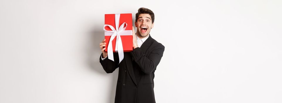 Image of happy good-looking man in black suit, holding box with christmas gift and smiling, standing against white background.