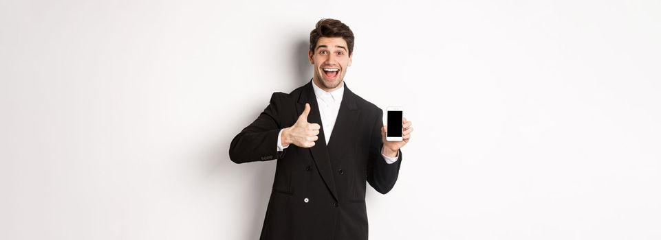 Image of handsome male entrepreneur in black suit, recommending app or online shop, showing thumbs-up and smartphone screen, standing over white background.