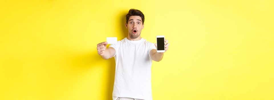 Handsome caucasian guy showing smartphone screen and credit card, concept of mobile banking and online shopping, yellow background.