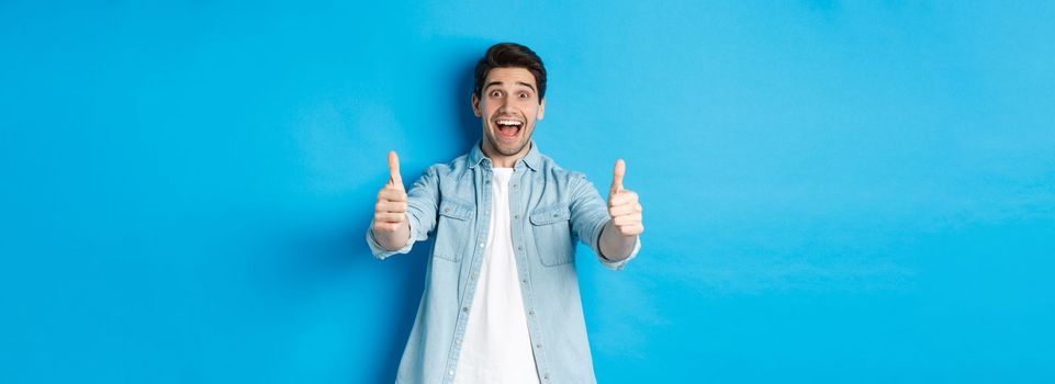Smiling confident man showing thumbs up with excited face, like something awesome, approving product, standing against blue background.