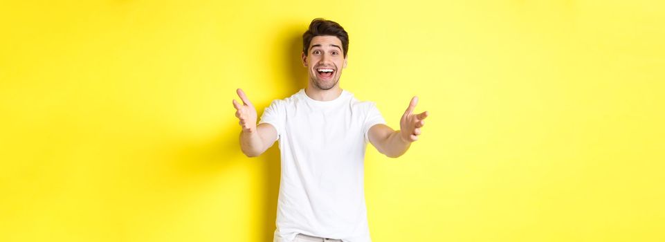 Excited handsome guy stretching hands forward, reaching for hug, receiving gift, standing over yellow background.