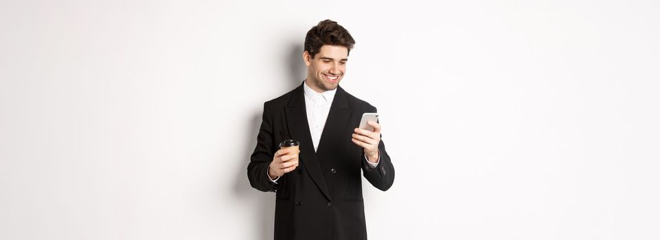 Portrait of handsome, confident businessman in black suit, drinking coffee and using mobile phone, smiling pleased, standing over white background.