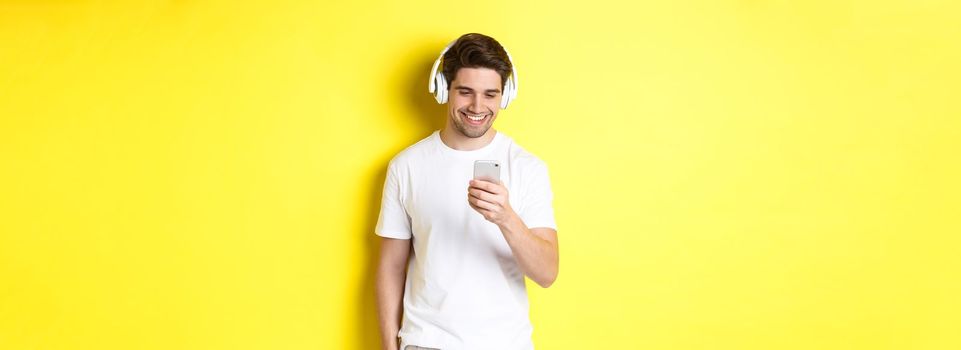 Young man in headphones reading message on smartphone, smiling, listening music in earphones and picking song in app, standing over yellow background.