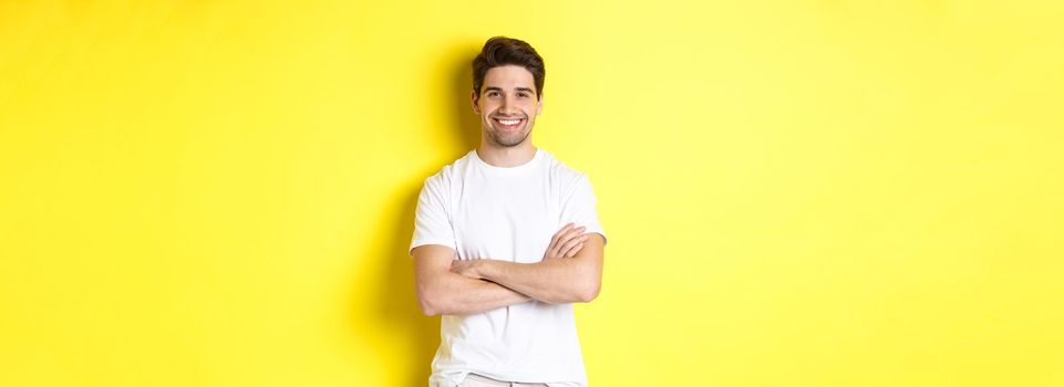 Image of confident caucasian man smiling pleased, holding hands crossed on chest and looking satisfied, standing over yellow background.